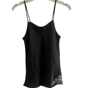 Svart linnetopp med detalj  - Snygg svart linnetopp med tunna axelband och dekorativ detalj i vitt längst ner på ena sidan. Toppens material känns mjukt och har en lätt glans.