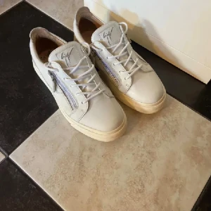 Vita sneakers från Giuseppe Zanotti - Snygga vita sneakers från Giuseppe Zanotti med signaturlogga på plösen och silverfärgade dragkedjor på sidorna. Skorna har snörning, rund tå och platt sula. Tillverkade i skinn för en lyxig känsla och stilren look.