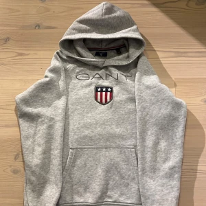 Grå hoodie från GANT med emblem - Snygg grå hoodie från GANT med broderat emblem och logga på bröstet. Tröjan har huva, lång ärm och en stor magficka framtill. Tillverkad i mjuk bomullsmix som är skön att ha på sig. Någon enstaka fläck på framfickan går säkert bort i tvätten 👍