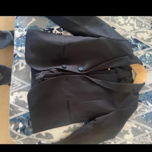 Svart klassisk kavaj med två knappar - Snygg svart kavaj med två knappar framtill och klassisk krage. Modellen har raka linjer och två fickor utan lock. Perfekt för dig som vill ha en stilren och tidlös look. Passar till skjorta eller t-shirt för en clean outfit.