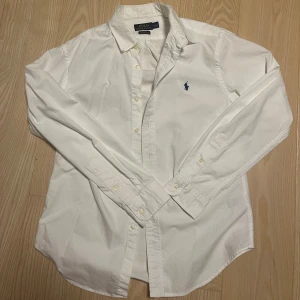 Vit slim fit skjorta från Polo Ralph Lauren - Vit skjorta från Polo Ralph Lauren i slim fit-modell, tillverkad i stretchig bomull. Klassisk oxford-krage och knappar framtill. Diskret blå broderad logga på bröstet. Perfekt för dig som gillar stilrena och tidlösa plagg.