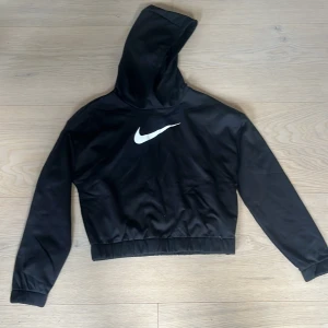 Nike hoodie Therma-FIT - Svart hoodie från Nike med Therma-FIT-teknologi och stor vit swoosh-logga på bröstet. Croppad modell med resår i midjan, lång ärm och huva. Perfekt för träning eller chill. Snygg och sportig stil som funkar året runt.          Aldrig använd 