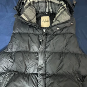 Svart pufferjacka från Burberry - Svart pufferjacka från Burberry armana finns också med klassiskt rutigt foder i huvan. Jackan har dragkedja och knappar framtill samt dragkedjeförsedda fickor. Materialet är polyester och dun, perfekt för kalla dagar. Stilsäker och tidlös design med märkets logga på detaljerna.