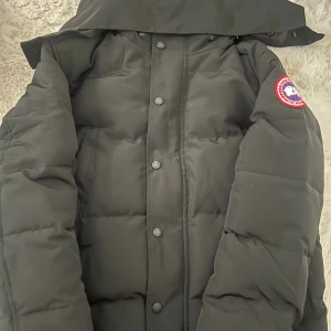 Svart dunjacka från Canada Goose XS - Säljer en svart dunjacka från Canada Goose i storlek XS. Jackan har huva, klassisk logga på ärmen och stängs med både dragkedja och knappar. Perfekt för kalla dagar med sin tjocka vaddering och stilrena look. Har fler storlekar.