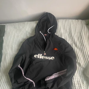 Svart vindjacka från Ellesse - Säljer en svart vindjacka från Ellesse med huva och vit dragsko. Jackan har halv dragkedja framtill, stor logga på bröstet och diskreta lila detaljer längs ärmarna. Perfekt för en sportig och avslappnad stil.