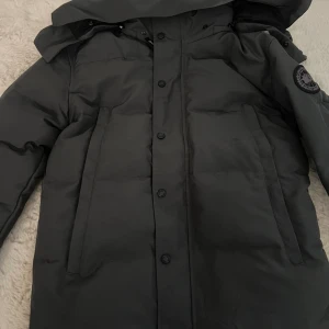 Svart dunjacka från Canada Goose XS - Säljer en svart dunjacka från Canada Goose i storlek XS. Jackan har huva, raka fickor och klassisk patch på ärmen. Perfekt för kalla dagar och har en stilren, enkel design med knappar framtill. Materialet är slitstarkt och håller värmen riktigt bra.