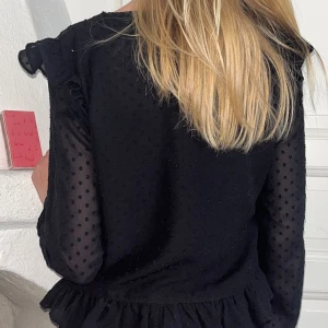 Svart prickig klänning med volang - Supersöt svart klänning med diskreta prickar, volangkant nertill och transparenta långärmar. V-ringad framtill med små knappar och fina detaljer längs sömmarna. Perfekt för dig som gillar en stilren men ändå lekfull look.