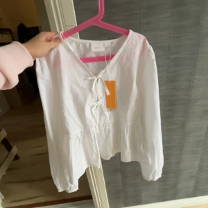 Vit blus från Gina tricot  - Säljer en vit blus med v-ringning och snörning framtill. Blusen har långa ärmar med resår vid ärmslut och en lätt volang nedtill. Perfekt för en fräsch och luftig look. Tillverkad i bomull för en skön känsla.