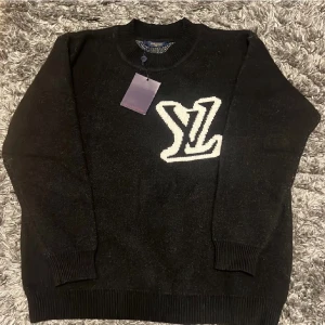 Svart stickad tröja Louis Vuitton  - Svart stickad tröja från Louis Vuitton i storlek S med stor vit LV-logga på bröstet. Rund halsringning och ribbade muddar vid ärmslut och nederkant. Perfekt för dig som gillar exklusiva märken och vill ha en stilren look.