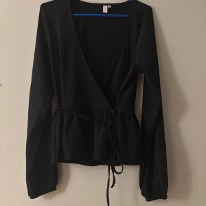 Svart omlottopp med knyt i midjan - Säljer en svart omlottopp från Gina Tricot med långa ärmar och v-ringad hals. Toppen har ett snyggt knyt i midjan och är tillverkad i ett mjukt, lätt strukturerat material. Perfekt för dig som gillar stilrena och trendiga plagg.