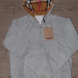 Grå hoodie från Burberry med dragkedja - Grå hoodie från Burberry med klassisk rutmönstrad huva i beige, svart och rött. Tröjan har dragkedja framtill, ribbade muddar och broderad Burberry-logga på bröstet. Perfekt för dig som gillar stilrena och exklusiva plagg med snygga detaljer.