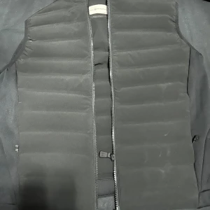 Svart väst från Peak Performance - Svart dunväst från Peak Performance med quiltad design och rak passform. Västen har en hel dragkedja framtill och är tillverkad i polyester med dunfyllning. Perfekt att slänga över en hoodie för extra värme. Snygg, stilren och enkel att matcha. Har använt en gång. Storlek L. Köpt på Hedes i Kungsbacka för 1000 har tyvärr inte kvar kvittot. Övriga frågor är det bara att skriva! 