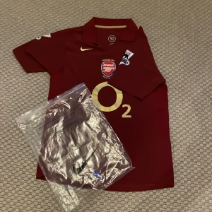Arsenal Highbury Bergkamp 10 Nike tröja - Exklusiv Arsenal Highbury 1913-2006 matchtröja i mörkröd med guldtryck, Bergkamp 10 på ryggen och O2-logga fram. Broderat klubbmärke, Premier League-patchar på ärmarna och klassisk krage. Tillverkad i lätt polyester, perfekt för samlare eller fans.