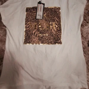 Vit t-shirt med leopardmönster från Guess - Snygg vit t-shirt från Guess med ett stort fyrkantigt leopardmönster framtill och guldfärgade detaljer. Trycket har texten 'Guess' i guld och 'Est. 1981'. T-shirten är kortärmad och i mjuk bomull, perfekt för en trendig look.
