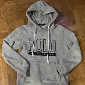 Grå hoodie från Polo Ralph Lauren - Snygg grå hoodie från Polo Ralph Lauren med stor logga framtill. Tröjan har huva med vita snören, magficka och ribbade muddar vid ärmslut och nederkant. Perfekt för chill dagar och streetstyle. Mjuk och skön bomullsmix som håller dig bekväm hela dagen.