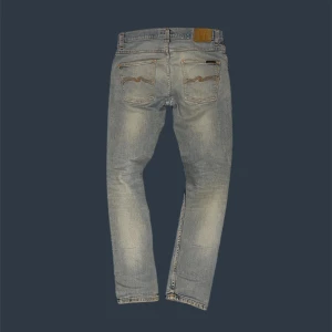 Nudie Jeans - Ljusblå jeans från Nudie Jeans med slitningar och lagade hål på knä och ficka. Modellen är Grim Tim. Längden är 108cm, midjan är 42cm, benöppning är 17cm, låret är 24cm.
