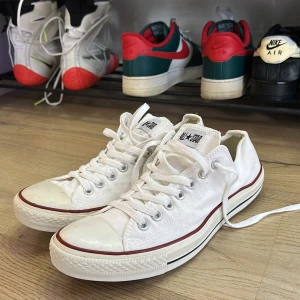 Vita Converse All Star sneakers - Säljer ett par klassiska vita Converse All Star sneakers med lågt skaft. Skorna har snörning, rund tå och platt sula med röd och svart rand längs kanten. Tillverkade i bomullstyg med gummidetaljer på tå och sula. Perfekta för dig som gillar tidlös streetstil.