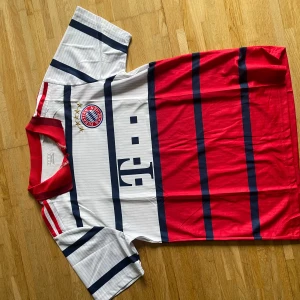 Bayern München randig fotbollströja - Snygg Bayern München fotbollströja från Adidas med breda röda och vita ränder, mörkblå detaljer och klubbmärke med fyra stjärnor på bröstet. Tröjan har korta ärmar, röd krage och är tillverkad i lätt polyester som andas. Perfekt för dig som älskar fotboll och vill sticka ut.