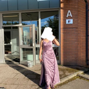Ljuslila långklänning med slits - Säljer en elegant ljuslila långklänning i glansigt material med omlottliv och smala axelband. Klänningen har en snygg slits framtill och markerad midja som ger en riktigt fin siluett. Perfekt för bal eller student!
