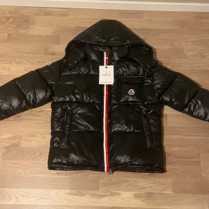Svart Moncler pufferjacka med huva - Svart pufferjacka från Moncler med glansig finish, huva och rödvit randig dragkedja. Jackan har en bröstficka med Moncler-logga, två sidofickor och coolt serietryck på insidan. Perfekt för kalla dagar och riktigt snygg streetstil.