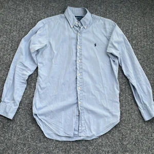 Ljusblå skjorta från Ralph Lauren - Klassisk ljusblå skjorta från Ralph Lauren med button-down krage och broderad marinblå logga på bröstet. Skjortan har lång ärm, normal passform och är tillverkad i mjuk bomull. Perfekt för en clean och stilren look. Den har en liten skavank på armbågen som man ser på sista bilden, och kanske också någon liten prick någon stans men inget som märks. Hör av dig vid frågor. Priser ej hugget i sten😊