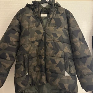 Mönstrad pufferjacka från Lindex - Säljer en pufferjacka från Lindex i storlek 170. Jackan har ett coolt geometriskt mönster i mörkgrön och grå ton, huva med ljusgrå insida och två fickor framtill. Vattenavvisande material, OBS HAR ETT LITET HÅL PÅ BAKSIDan