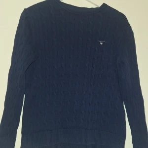 Marinblå kabelstickad tröja från Gant - Kabelstickad marinblå tröja från Gant med broderad logga på bröstet. Tröjan har rund halsringning, ribbade muddar och är långärmad. Perfekt för dig som gillar klassisk och stilren look.