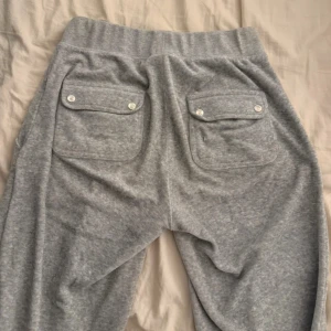 Grå mjukisbyxor Juicy Couture XS - Grå mjukisbyxor från Juicy Couture med broderad JC-logga på fickan. Byxorna har bred resår i midjan, snörning och två bakfickor med lock och silvriga knappar. Mjukt bomullsmaterial som är perfekt för chill dagar.