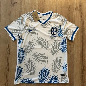 Vit Brasilien fotbollströja Nike M - Snygg Brasilien fotbollströja från Nike i vitt med blå och grå palmbladsmönster. Kortärmad med blå detaljer vid krage och ärmslut, samt CBF-emblem på bröstet. Tillverkad i Dri-FIT material som andas och håller dig sval under matchen.