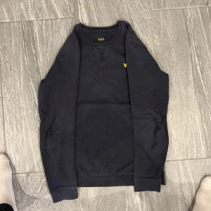 Mörkblå sweatshirt från Lyle & Scott - Säljer en stilren mörkblå sweatshirt från Lyle & Scott med klassisk rund hals och det ikoniska gula fågel-loggan broderad på bröstet. Tröjan har långa ärmar och är tillverkad i mjuk bomull, perfekt för en chill och snygg look.
