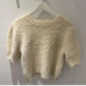 Vit fluffig stickad croppad tröja - Supermysig vit stickad tröja med korta ärmar och croppad passform. Tröjan har en rund halsringning och är gjord i ett fluffigt material som känns mjukt mot huden. Perfekt för dig som gillar en chill och trendig look.