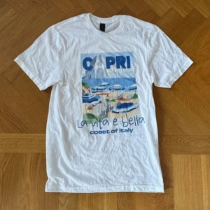 Vit Capri t-shirt med tryck - Vit t-shirt med färgglatt tryck av Capri och texten 'La vita e bella, coast of Italy'. Motiv med strand, parasoller och hav i blå toner. Klassisk passform och rund halsringning. Perfekt för dig som gillar sommarvibbar och italiensk stil.