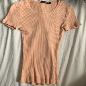 Ribbad persikofärgad topp från Bik Bok - Säljer en ribbad, persikofärgad topp från Bik Bok i storlek S. Toppen har rund halsringning, korta ärmar och är figurnära i modellen. Perfekt att styla med jeans eller kjol för en enkel och snygg look.