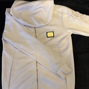 Vit hoodiejacka från Black Squad - Snygg vit hoodiejacka från Black Squad med guldfärgad dragkedja och detaljer. Jackan har huva, ribbade muddar och ett guldmärke på bröstet. På ena ärmen står det 'BLACK SQUAD' i relief. Perfekt för dig som gillar streetwear och vill sticka ut.