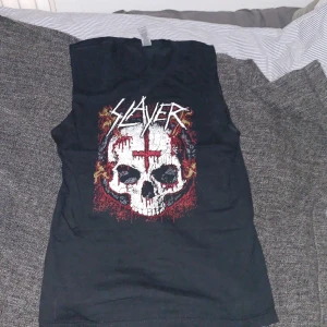 Svart Slayer ärmlös t-shirt - Säljer en svart ärmlös t-shirt med Slayer-tryck framtill. Stort motiv med dödskalle, röd kors och eldiga detaljer. Perfekt för dig som gillar metal och vill sticka ut med en cool bandtröja.