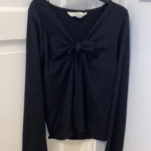 Svart långärmad topp med rosett H&M - Svart långärmad topp från H&M med snygg v-ringning och stor rosett framtill. Mjuk och stretchig bomullskvalitet som sitter skönt. Perfekt för dig som gillar stilrena detaljer och vill ha en enkel men ändå cool look.