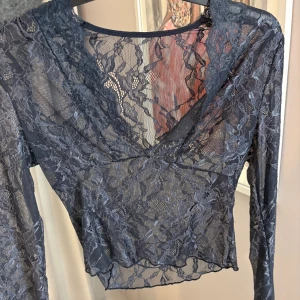 Mörkblå spetsblus med v-ringning - Säljer en mörkblå, transparent spetsblus med v-ringad hals och långa ärmar. Blusen har ett blommigt spetsmönster och är croppad i modellen. Perfekt för dig som vill ha en snygg och trendig topp med lite edge.