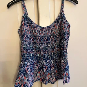 Mönstrad blå linnetopp från H&M Divided - Säljer en färgglad linnetopp från H&M Divided i storlek 36. Toppen har smala axelband, smockad överdel och ett coolt geometriskt mönster i blått, rosa och vitt. Perfekt för varma dagar och har en lätt, flowy känsla.