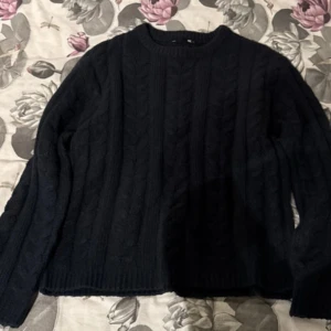 Mörkblå kabelstickad tröja - Mysig svart kabelstickad tröja med rund hals och långa ärmar. Tröjan har ett klassiskt flätmönster och ribbade muddar vid ärmslut och nederkant. Perfekt för kyliga dagar när du vill vara både bekväm och snygg.