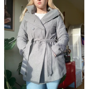 Grå kappa med huva och bälte - Snygg grå kappa med stor huva och knytskärp i midjan. Kappan har dubbelknäppning framtill, långa ärmar och rak passform. 