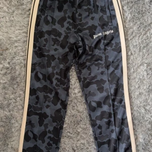 Palm Angels camo trackpants L - Palm Angels trackpants i storlek L med svart och grått camouflage-mönster, beige sidorand och logotyp på låret. Byxorna är gjorda i 100% polyester och har en modern, loose passform. Perfekt för dig som gillar streetwear och vill sticka ut.