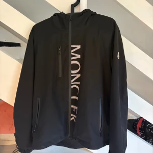 Svart vindjacka från Moncler med huva - Svart vindjacka från Moncler med stor logga framtill, huva och dragkedja. Jackan har tre fickor med dragkedja och Monclers klassiska röd-vit-blå detalj bak. Perfekt för dig som gillar stilren och sportig look.