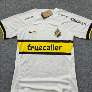 AIK vit bortatröja Nike Dri-FIT M - AIK:s officiella vita bortatröja från Nike i Dri-FIT-material. Tröjan har korta ärmar, svart och gul rand över bröstet med truecaller-tryck, AIK-emblem och Nike-logga. Stadium, Craftor och SVEA-loggor på ärmar och rygg. Perfekt för dig som vill visa ditt AIK-stöd.
