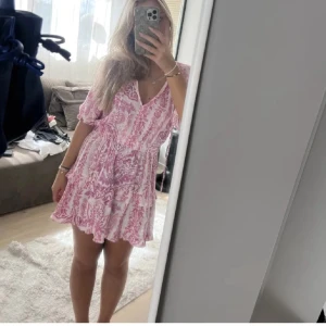 Rosa och vit kortklänning med volang - Superfin kortklänning i vitt och rosa med blommigt mönster. Klänningen har v-ringning med knyt, volangkant nertill och resår i midjan. Lätt och luftig med korta ärmar, perfekt för varma dagar.