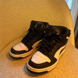 Svarta och vita sneakers från Puma storlek 38 - Snygga high-top sneakers från Puma i svart och vitt med klassisk snörning. Skorna har perforerad tå, rund form och en platt sula. Materialet är skinn och syntet, vilket ger en cool och sportig vibe. Perfekt för dig som gillar streetstyle och vill sticka ut.