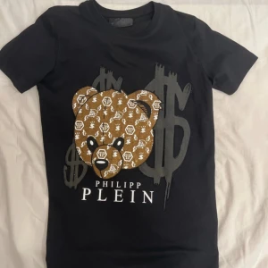 Svart Philipp Plein t-shirt med björn - Svart t-shirt från Philipp Plein med korta ärmar och normal passform. Framsidan har en stor björn i brunt med vita loggor och svarta detaljer, samt stora grå dollarsymboler i bakgrunden. Materialet är mjuk bomull och trycket är detaljerat och sticker ut.