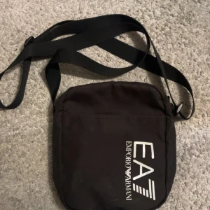 Svart axelväska från Emporio Armani - Svart axelväska från Emporio Armani EA7 med justerbar rem och vit logga på framsidan. Väskan har dragkedja upptill och 3 fack inuti för smidig organisering och en utanför. Perfekt storlek för mobil, plånbok och andra små och stora saker. Snygg och stilren design.