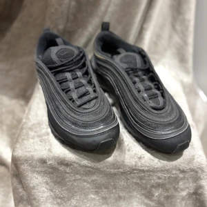 Nike Air Max 97 Triple Black 42 - Säljer ett par Nike Air Max 97 i helsvart färg. Skorna har den ikoniska vågiga designen, synlig Air Max-sula och snörning. Tillverkade i en mix av syntet och textil med detaljer i skinn. Perfekta för dig som gillar en clean och sportig look.