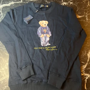 Mörkblå Polo Bear tröja Ralph Lauren - Snygg mörkblå tröja från Ralph Lauren med ikoniska Polo Bear-trycket framtill. Tröjan har rund halsringning, långa ärmar och ribbade muddar. Tillverkad i mjuk bomull som är skön att ha på sig. Perfekt för dig som gillar streetstyle och klassiska märken.