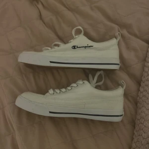 Vita Champion sneakers med snören - Snygga vita sneakers från Champion med klassisk låg profil och rund tå. Skorna har vita snören och är tänkt att använda inomhus, därav kan jag inte garantera att de är vattentäta. Champion-loggan är broderad i svart på sidan. Yttersulan är gummibrun. Använda 1 gång. Vid frågor eller fler bilder, kontakta mig. 